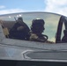 A military pilot is seated in the cockpit of a modern stealth fighter aircraft, identifiable by its low-observable canopy design and warning markings. The pilot is wearing a full flight helmet with oxygen mask and flight suit. The image is taken from outside the closed canopy, looking in at an angle against a partly cloudy sky.