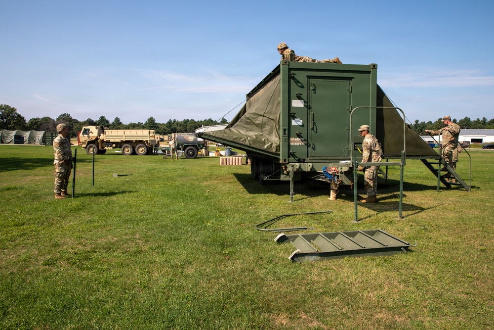 Military personnel in camouflage uniforms are setting up or breaking down a large green military field shelter or expandable container unit on a grassy field. Several soldiers are working on the structure, with one on top and others on stairs and ground level. Additional military vehicles and equipment are visible in the background.