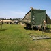 Military personnel in camouflage uniforms are setting up or breaking down a large green military field shelter or expandable container unit on a grassy field. Several soldiers are working on the structure, with one on top and others on stairs and ground level. Additional military vehicles and equipment are visible in the background.