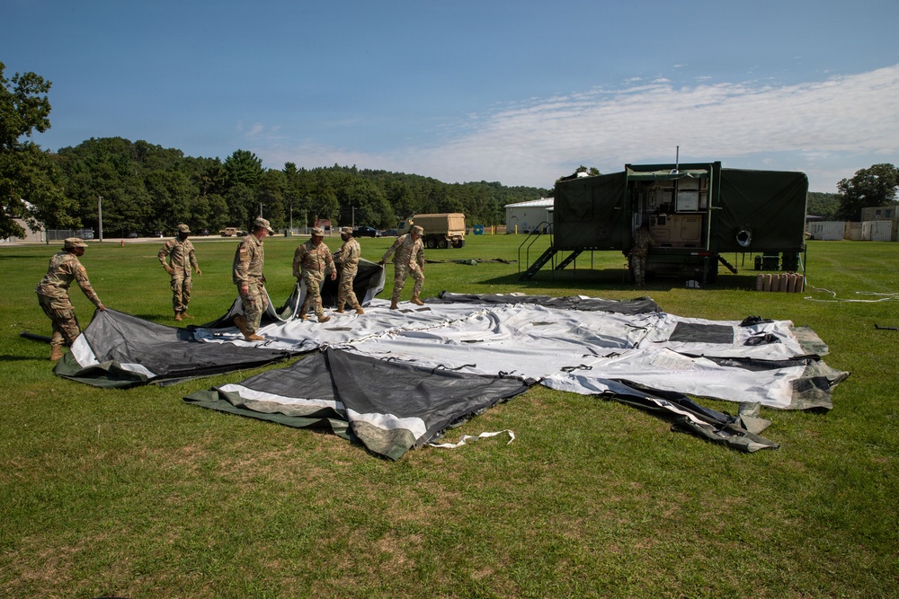 A group of military personnel in camouflage uniforms are working together on a grassy field to lay out or set up a large tent or tarpaulin structure. The scene appears to be at a military installation or training area, with military vehicles and a large green shelter unit visible in the background. The activity suggests field operations setup or training exercises.