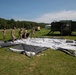 A group of military personnel in camouflage uniforms are working together on a grassy field to lay out or set up a large tent or tarpaulin structure. The scene appears to be at a military installation or training area, with military vehicles and a large green shelter unit visible in the background. The activity suggests field operations setup or training exercises.