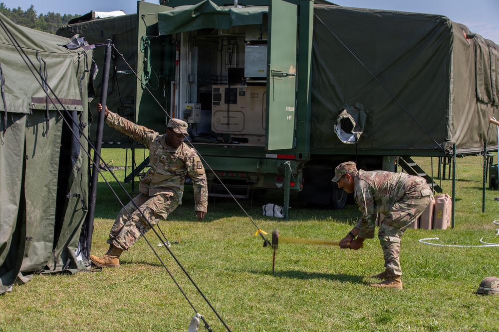 Two U.S. Army soldiers in MultiCam/OCP camouflage uniforms are setting up a military field camp, with one soldier holding a tent pole and the other driving a ground stake with a mallet. Behind them is a large green military shelter/command post vehicle with its rear panel open, revealing electronic equipment inside. The scene appears to be a field training exercise or operational deployment on a grassy area.