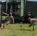 Two U.S. Army soldiers in MultiCam/OCP camouflage uniforms are setting up a military field camp, with one soldier holding a tent pole and the other driving a ground stake with a mallet. Behind them is a large green military shelter/command post vehicle with its rear panel open, revealing electronic equipment inside. The scene appears to be a field training exercise or operational deployment on a grassy area.