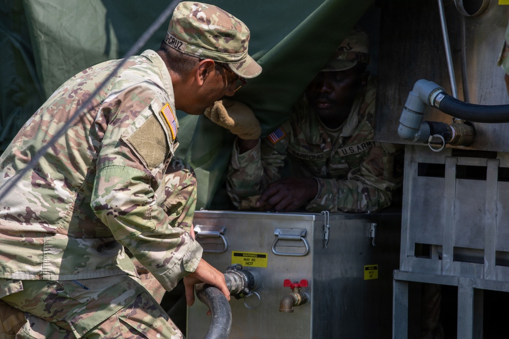 Two U.S. Army soldiers in OCP (Operational Camouflage Pattern) uniforms are working with what appears to be a field water purification or heating unit. One soldier is leaning over the stainless steel equipment while the other is positioned inside or behind the unit. The setting appears to be a field environment with a military tent visible in the background.