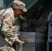Two U.S. Army soldiers in OCP (Operational Camouflage Pattern) uniforms are working with what appears to be a field water purification or heating unit. One soldier is leaning over the stainless steel equipment while the other is positioned inside or behind the unit. The setting appears to be a field environment with a military tent visible in the background.