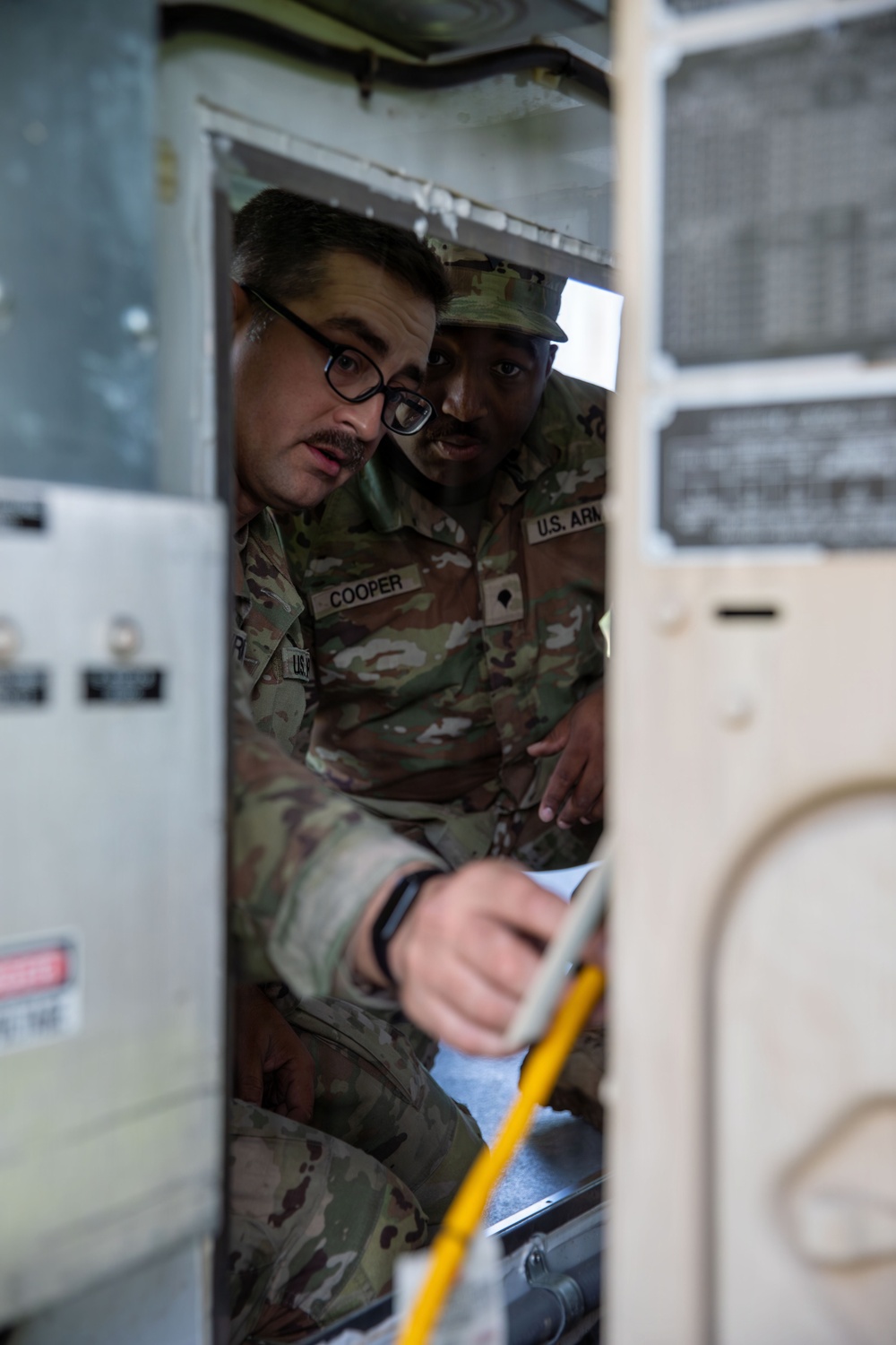 Two U.S. Army soldiers in OCP (Operational Camouflage Pattern) uniforms are examining or working inside what appears to be a military vehicle or equipment compartment. One soldier, identified by a name tape as 'COOPER,' is visible along with a second soldier wearing glasses. They appear to be conducting maintenance, inspection, or training on the equipment.