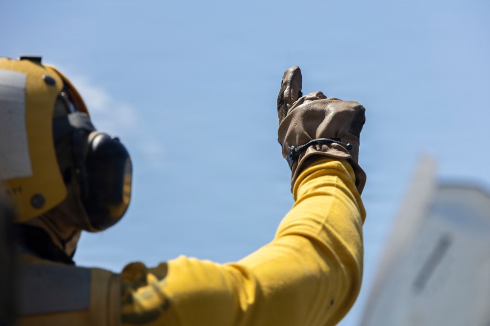 A person wearing a yellow flight deck jersey and brown leather gloves is giving a thumbs-up signal against a clear blue sky. The individual is also wearing a yellow helmet equipped with hearing protection/communication ear cups. This appears to be a naval aircraft carrier flight deck crew member performing a standard hand signal.