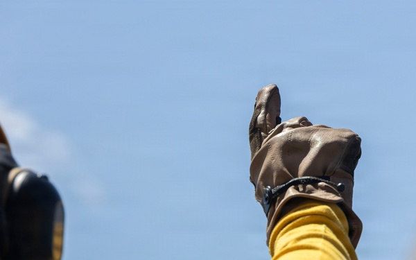 A person wearing a yellow flight deck jersey and brown leather gloves is giving a thumbs-up signal against a clear blue sky. The individual is also wearing a yellow helmet equipped with hearing protection/communication ear cups. This appears to be a naval aircraft carrier flight deck crew member performing a standard hand signal.