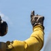 A person wearing a yellow flight deck jersey and brown leather gloves is giving a thumbs-up signal against a clear blue sky. The individual is also wearing a yellow helmet equipped with hearing protection/communication ear cups. This appears to be a naval aircraft carrier flight deck crew member performing a standard hand signal.