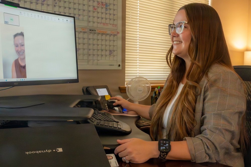 A young woman with long auburn hair and glasses sits at an office desk engaged in a video call on an AOC desktop monitor. She is smiling and appears to be in a professional office environment with a wall calendar, desk phone, and small fan visible in the background. A Dynabook laptop is also present on the desk in front of her.