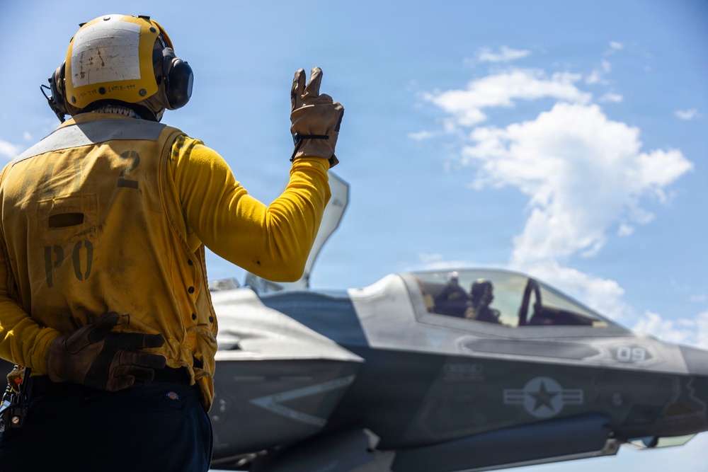 A U.S. Navy flight deck crew member in a yellow jersey is signaling to an F-35 fighter jet on what appears to be an aircraft carrier flight deck. The deck handler, wearing a yellow helmet and gloves, is giving a hand signal with two fingers raised toward the aircraft. The F-35, bearing the number '09' and a U.S. military star insignia, has a pilot visible in the cockpit.