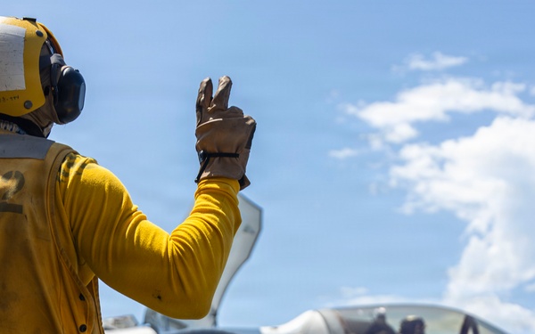 A U.S. Navy flight deck crew member in a yellow jersey is signaling to an F-35 fighter jet on what appears to be an aircraft carrier flight deck. The deck handler, wearing a yellow helmet and gloves, is giving a hand signal with two fingers raised toward the aircraft. The F-35, bearing the number '09' and a U.S. military star insignia, has a pilot visible in the cockpit.