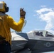 A U.S. Navy flight deck crew member in a yellow jersey is signaling to an F-35 fighter jet on what appears to be an aircraft carrier flight deck. The deck handler, wearing a yellow helmet and gloves, is giving a hand signal with two fingers raised toward the aircraft. The F-35, bearing the number '09' and a U.S. military star insignia, has a pilot visible in the cockpit.