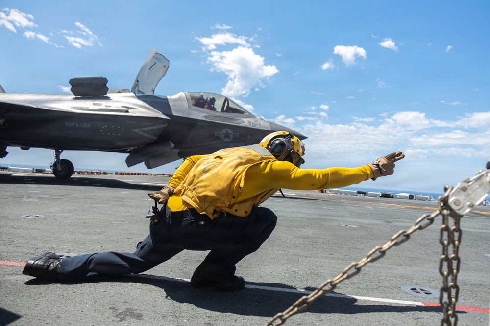 A U.S. Navy flight deck crew member wearing a yellow jersey is performing a launch signal gesture in a crouched position on an aircraft carrier flight deck. Behind them is an F-35B Lightning II stealth fighter jet marked with 'VMFA-214' (Marine Fighter Attack Squadron 214) and the number '09'. The scene takes place on an open ocean carrier deck under a partly cloudy blue sky.