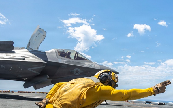 A U.S. Navy flight deck crew member wearing a yellow jersey is performing a launch signal gesture in a crouched position on an aircraft carrier flight deck. Behind them is an F-35B Lightning II stealth fighter jet marked with 'VMFA-214' (Marine Fighter Attack Squadron 214) and the number '09'. The scene takes place on an open ocean carrier deck under a partly cloudy blue sky.