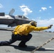 A U.S. Navy flight deck crew member wearing a yellow jersey is performing a launch signal gesture in a crouched position on an aircraft carrier flight deck. Behind them is an F-35B Lightning II stealth fighter jet marked with 'VMFA-214' (Marine Fighter Attack Squadron 214) and the number '09'. The scene takes place on an open ocean carrier deck under a partly cloudy blue sky.