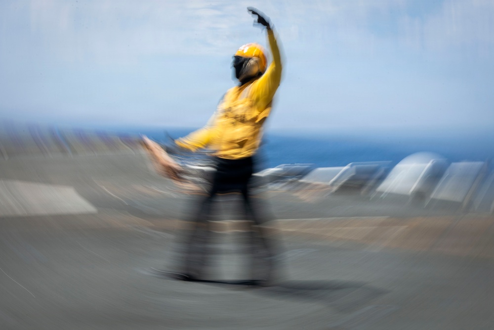 A motion-blurred photograph captures a flight deck crew member aboard what appears to be an aircraft carrier, wearing a yellow jersey and helmet while performing a launch signal gesture. The individual has one arm raised overhead in a characteristic aircraft launch or directing signal. The image was taken with intentional or incidental camera motion, creating a radial blur effect around the subject.
