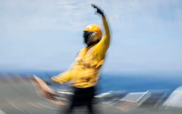 A motion-blurred photograph captures a flight deck crew member aboard what appears to be an aircraft carrier, wearing a yellow jersey and helmet while performing a launch signal gesture. The individual has one arm raised overhead in a characteristic aircraft launch or directing signal. The image was taken with intentional or incidental camera motion, creating a radial blur effect around the subject.