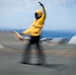 A motion-blurred photograph captures a flight deck crew member aboard what appears to be an aircraft carrier, wearing a yellow jersey and helmet while performing a launch signal gesture. The individual has one arm raised overhead in a characteristic aircraft launch or directing signal. The image was taken with intentional or incidental camera motion, creating a radial blur effect around the subject.