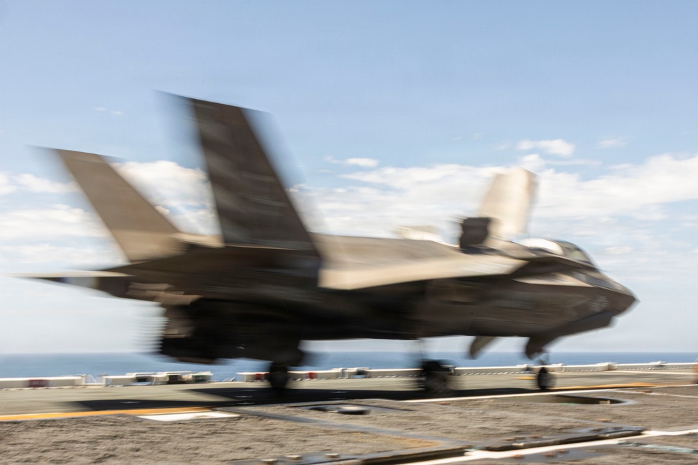 A military stealth fighter jet, consistent with an F-35B variant, is captured in motion on what appears to be the flight deck of an aircraft carrier or amphibious assault ship. The image exhibits significant motion blur, indicating the aircraft is moving at high speed during takeoff or landing. The ocean horizon is visible in the background under a partly cloudy sky.