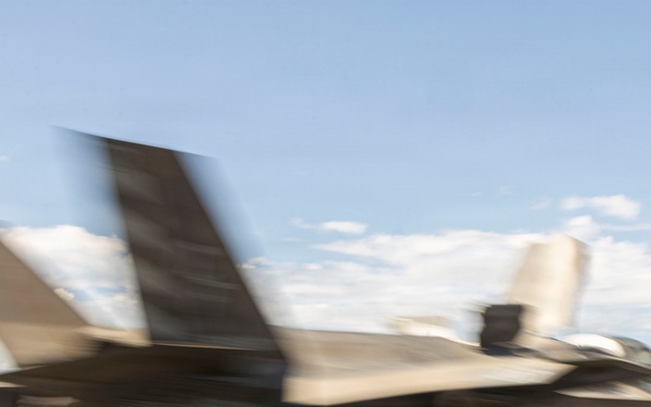 A military stealth fighter jet, consistent with an F-35B variant, is captured in motion on what appears to be the flight deck of an aircraft carrier or amphibious assault ship. The image exhibits significant motion blur, indicating the aircraft is moving at high speed during takeoff or landing. The ocean horizon is visible in the background under a partly cloudy sky.