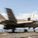 A military stealth fighter jet, consistent with an F-35B variant, is captured in motion on what appears to be the flight deck of an aircraft carrier or amphibious assault ship. The image exhibits significant motion blur, indicating the aircraft is moving at high speed during takeoff or landing. The ocean horizon is visible in the background under a partly cloudy sky.