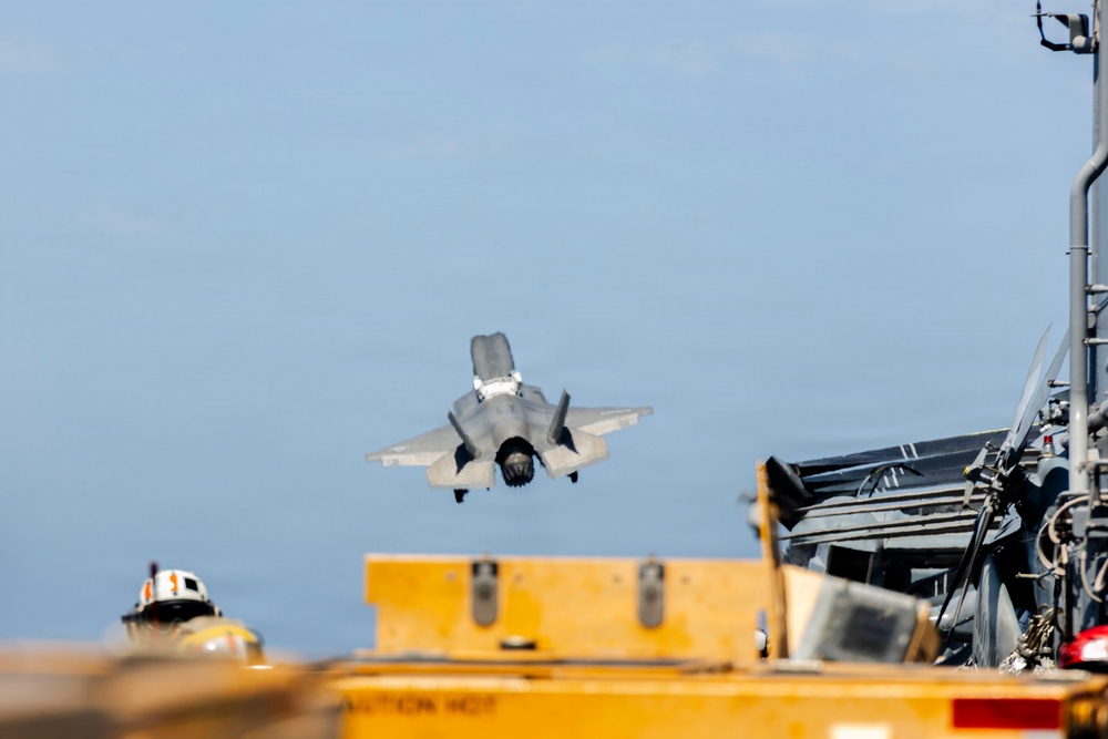 A fifth-generation stealth fighter jet, consistent with an F-35B, is captured in a vertical or short takeoff/landing (VSTOL) maneuver above what appears to be the deck of an amphibious assault ship or aircraft carrier. The aircraft's landing gear is deployed and it is hovering or in very low-speed flight close to the ship's deck. Deck crew personnel in flight deck gear are visible in the foreground.