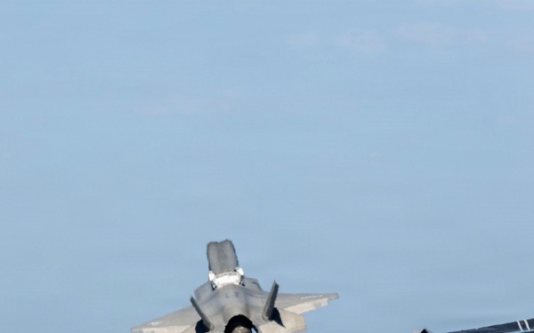A fifth-generation stealth fighter jet, consistent with an F-35B, is captured in a vertical or short takeoff/landing (VSTOL) maneuver above what appears to be the deck of an amphibious assault ship or aircraft carrier. The aircraft's landing gear is deployed and it is hovering or in very low-speed flight close to the ship's deck. Deck crew personnel in flight deck gear are visible in the foreground.