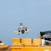 A fifth-generation stealth fighter jet, consistent with an F-35B, is captured in a vertical or short takeoff/landing (VSTOL) maneuver above what appears to be the deck of an amphibious assault ship or aircraft carrier. The aircraft's landing gear is deployed and it is hovering or in very low-speed flight close to the ship's deck. Deck crew personnel in flight deck gear are visible in the foreground.