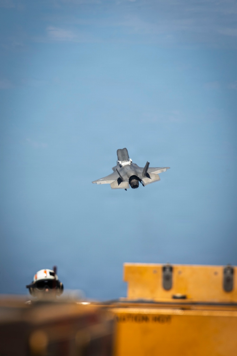 A fifth-generation stealth fighter jet, consistent with an F-35 variant, is captured in flight against a clear blue sky. The aircraft appears to be in a near-vertical climb or steep banking maneuver, viewed from a head-on/slightly below angle. In the foreground, partially out of focus, are yellow military ground support equipment containers and a person wearing a white helmet, suggesting a naval or military flight deck environment.