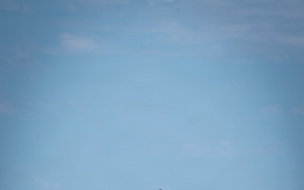 A fifth-generation stealth fighter jet, consistent with an F-35 variant, is captured in flight against a clear blue sky. The aircraft appears to be in a near-vertical climb or steep banking maneuver, viewed from a head-on/slightly below angle. In the foreground, partially out of focus, are yellow military ground support equipment containers and a person wearing a white helmet, suggesting a naval or military flight deck environment.
