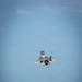 A fifth-generation stealth fighter jet, consistent with an F-35 variant, is captured in flight against a clear blue sky. The aircraft appears to be in a near-vertical climb or steep banking maneuver, viewed from a head-on/slightly below angle. In the foreground, partially out of focus, are yellow military ground support equipment containers and a person wearing a white helmet, suggesting a naval or military flight deck environment.