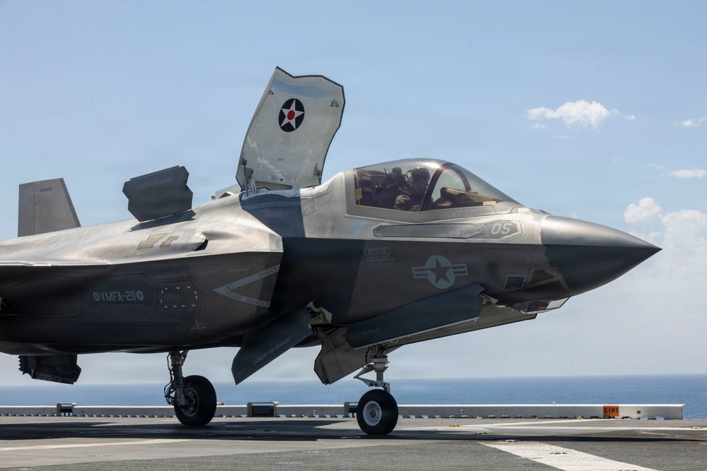 A U.S. Marine Corps F-35B Lightning II stealth fighter jet is positioned on the flight deck of an amphibious assault ship, with its lift fan door open indicating STOVL (Short Take-Off and Vertical Landing) capability. The aircraft bears markings from VMFA-211 (Marine Fighter Attack Squadron 211) and displays the number '05' on its fuselage. A pilot is visible in the cockpit, and the ocean horizon is visible in the background.