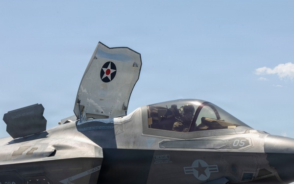 A U.S. Marine Corps F-35B Lightning II stealth fighter jet is positioned on the flight deck of an amphibious assault ship, with its lift fan door open indicating STOVL (Short Take-Off and Vertical Landing) capability. The aircraft bears markings from VMFA-211 (Marine Fighter Attack Squadron 211) and displays the number '05' on its fuselage. A pilot is visible in the cockpit, and the ocean horizon is visible in the background.
