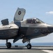 A U.S. Marine Corps F-35B Lightning II stealth fighter jet is positioned on the flight deck of an amphibious assault ship, with its lift fan door open indicating STOVL (Short Take-Off and Vertical Landing) capability. The aircraft bears markings from VMFA-211 (Marine Fighter Attack Squadron 211) and displays the number '05' on its fuselage. A pilot is visible in the cockpit, and the ocean horizon is visible in the background.