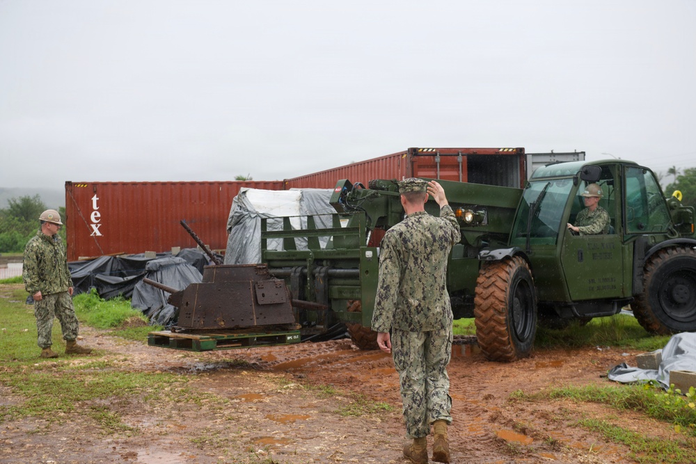 Military personnel in Navy Working Uniform (NWU) camouflage are conducting a logistics or equipment handling operation in a muddy outdoor area. A green military rough terrain forklift (telehandler) is being directed by a service member acting as a ground guide. The scene includes shipping containers, palletized equipment, and what appears to be rusted or salvaged heavy machinery.