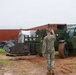 Military personnel in Navy Working Uniform (NWU) camouflage are conducting a logistics or equipment handling operation in a muddy outdoor area. A green military rough terrain forklift (telehandler) is being directed by a service member acting as a ground guide. The scene includes shipping containers, palletized equipment, and what appears to be rusted or salvaged heavy machinery.