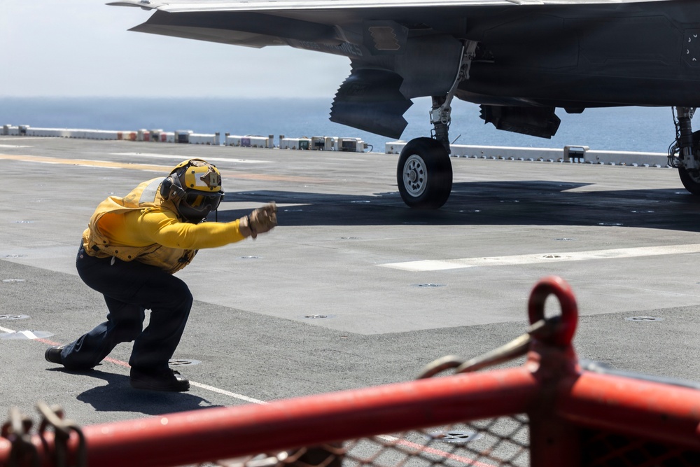 A naval flight deck crew member wearing a yellow jersey and helmet is giving a launch signal in a crouched position on an aircraft carrier flight deck. A military aircraft, identifiable by its stealth-shaped underside and landing gear, is positioned nearby ready for launch. Red safety barriers and deck equipment are visible in the foreground.