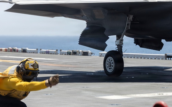 A naval flight deck crew member wearing a yellow jersey and helmet is giving a launch signal in a crouched position on an aircraft carrier flight deck. A military aircraft, identifiable by its stealth-shaped underside and landing gear, is positioned nearby ready for launch. Red safety barriers and deck equipment are visible in the foreground.