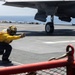 A naval flight deck crew member wearing a yellow jersey and helmet is giving a launch signal in a crouched position on an aircraft carrier flight deck. A military aircraft, identifiable by its stealth-shaped underside and landing gear, is positioned nearby ready for launch. Red safety barriers and deck equipment are visible in the foreground.