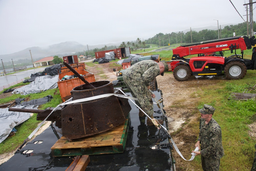 Two U.S. Navy personnel in camouflage uniforms are working outdoors in rainy conditions, handling straps or rigging around a heavily corroded, rusty metal object that appears to be a historical tank turret or gun emplacement on wooden pallets. The scene is set in what appears to be a military storage or staging yard with shipping containers, tarps, and a red XCMG telescopic forklift in the background. The environment suggests a tropical location, likely a Pacific island military installation.