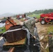 Two U.S. Navy personnel in camouflage uniforms are working outdoors in rainy conditions, handling straps or rigging around a heavily corroded, rusty metal object that appears to be a historical tank turret or gun emplacement on wooden pallets. The scene is set in what appears to be a military storage or staging yard with shipping containers, tarps, and a red XCMG telescopic forklift in the background. The environment suggests a tropical location, likely a Pacific island military installation.