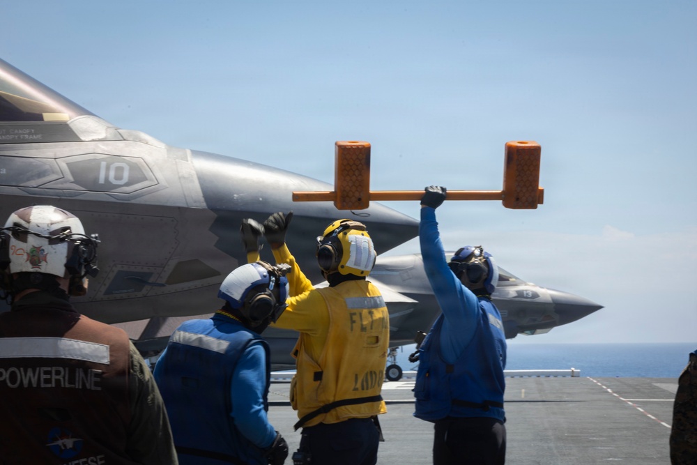 Flight deck crew personnel are directing aircraft operations aboard what appears to be a naval aircraft carrier. At least two F-35 stealth fighter jets are visible on the flight deck, numbered '10' and '13'. Deck crew in color-coded vests and helmets are using hand signals and a chock bar to coordinate aircraft movement.