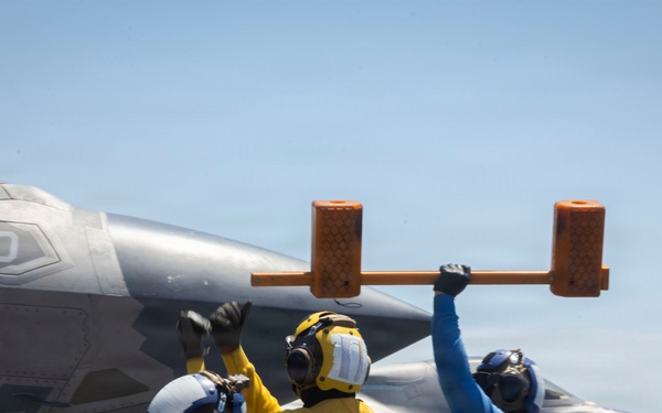 Flight deck crew personnel are directing aircraft operations aboard what appears to be a naval aircraft carrier. At least two F-35 stealth fighter jets are visible on the flight deck, numbered '10' and '13'. Deck crew in color-coded vests and helmets are using hand signals and a chock bar to coordinate aircraft movement.