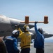 Flight deck crew personnel are directing aircraft operations aboard what appears to be a naval aircraft carrier. At least two F-35 stealth fighter jets are visible on the flight deck, numbered '10' and '13'. Deck crew in color-coded vests and helmets are using hand signals and a chock bar to coordinate aircraft movement.