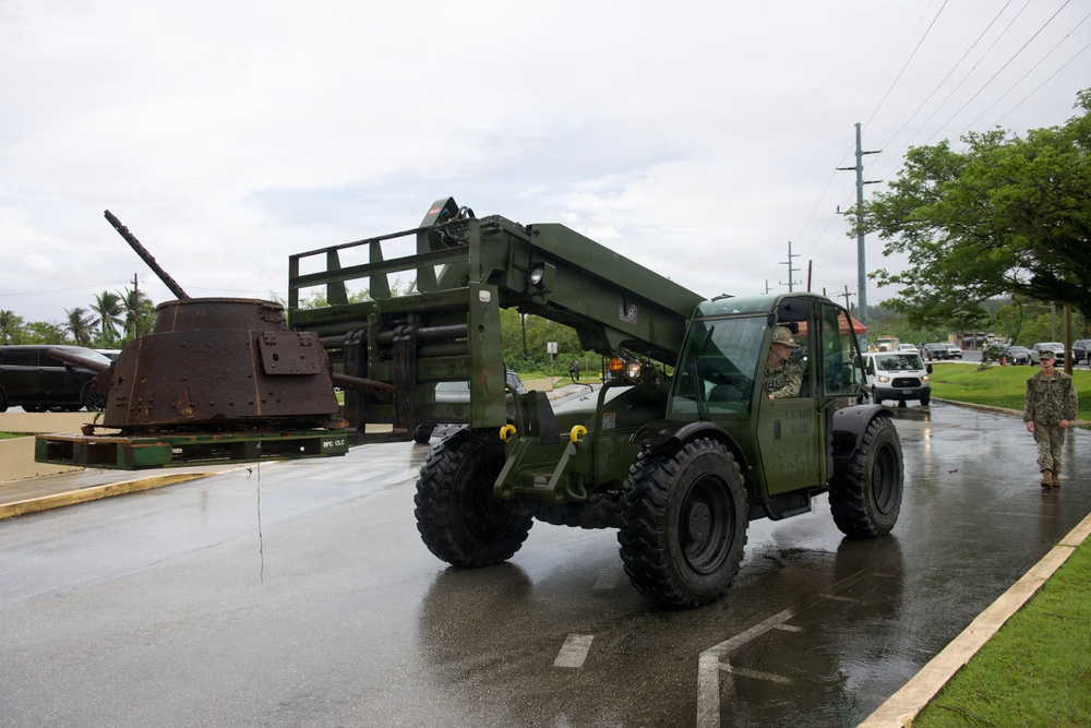 A military telehandler/rough terrain forklift, painted olive green and marked 'U.S. Navy,' is being operated on a wet paved surface near what appears to be a military base or installation. In the background, a heavily rusted historical artillery piece or tank turret is displayed on a wooden pallet, likely as a static museum exhibit. Two military personnel in Navy working uniforms are visible in the scene.
