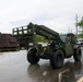 A military telehandler/rough terrain forklift, painted olive green and marked 'U.S. Navy,' is being operated on a wet paved surface near what appears to be a military base or installation. In the background, a heavily rusted historical artillery piece or tank turret is displayed on a wooden pallet, likely as a static museum exhibit. Two military personnel in Navy working uniforms are visible in the scene.