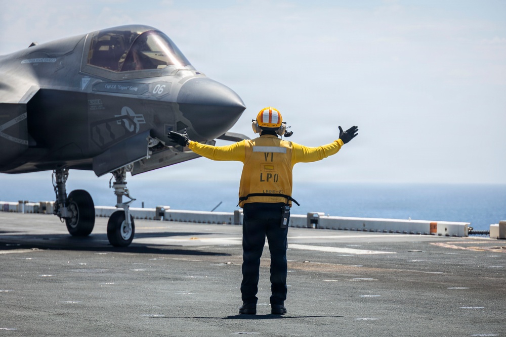 A flight deck crew member in a yellow jersey and orange helmet directs an F-35B stealth fighter jet numbered '06' on what appears to be the flight deck of a U.S. Navy amphibious assault ship. The crew member, identified by their vest as 'LPO' (Leading Petty Officer), has both arms extended in a signaling gesture. The ocean is visible in the background, confirming the shipboard setting.