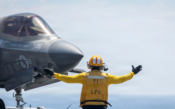A flight deck crew member in a yellow jersey and orange helmet directs an F-35B stealth fighter jet numbered '06' on what appears to be the flight deck of a U.S. Navy amphibious assault ship. The crew member, identified by their vest as 'LPO' (Leading Petty Officer), has both arms extended in a signaling gesture. The ocean is visible in the background, confirming the shipboard setting.
