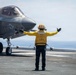 A flight deck crew member in a yellow jersey and orange helmet directs an F-35B stealth fighter jet numbered '06' on what appears to be the flight deck of a U.S. Navy amphibious assault ship. The crew member, identified by their vest as 'LPO' (Leading Petty Officer), has both arms extended in a signaling gesture. The ocean is visible in the background, confirming the shipboard setting.